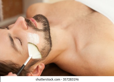 Close up of hand of beautician applying cream on male face with a brush. The man is lying and relaxing at spa