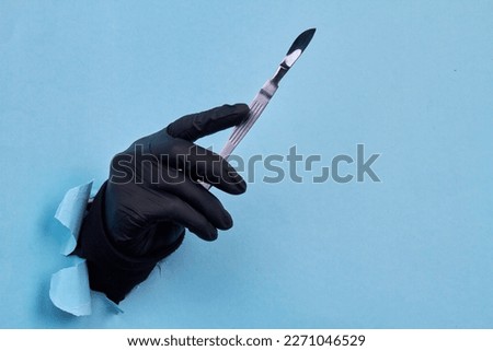 Close up hand in black glove holding scalpel. Blue paper background with copy space.