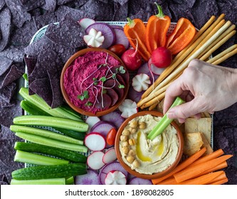 Close up of a hand dipping a celery stick into hummus dip with colourful vegetables and blue corn chips surrounding. - Powered by Shutterstock