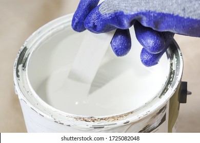 Close Up Of Hand Gloved Painter Mixing Paint In Metal Paint Can With Wooden Stick.