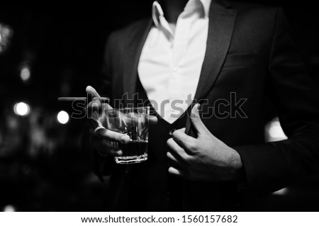 Close up hand of handsome well-dressed arabian man with glass of whiskey and cigar posed at pub.
