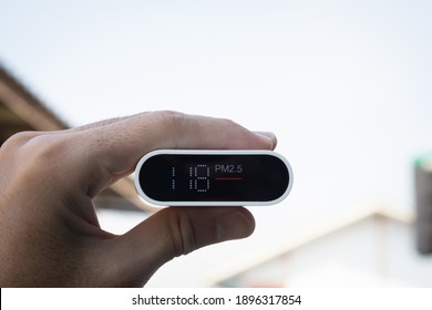 Close Up Hand Holding Air Quality Monitor To Detect Level Of Pollution Or Small Particle (PM2.5) On Home Background. Image High Pollution Concept.