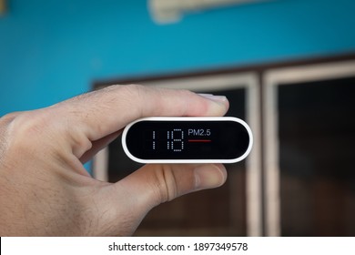 Close Up Hand Holding Air Quality Monitor To Detect Level Of Pollution Or Small Particle (PM2.5) On Home Background. Image High Pollution Concept.