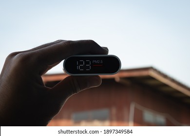 Close Up Hand Holding Air Quality Monitor To Detect Level Of Pollution Or Small Particle (PM2.5) On Home Background. Image High Pollution Concept.