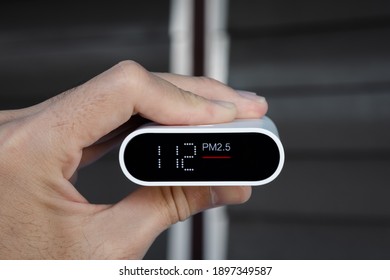 Close Up Hand Holding Air Quality Monitor To Detect Level Of Pollution Or Small Particle (PM2.5) On Home Background. Image High Pollution Concept.