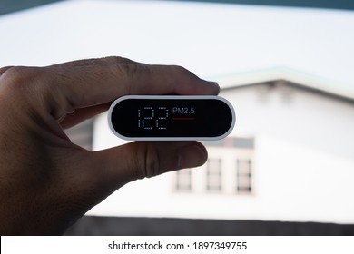 Close Up Hand Holding Air Quality Monitor To Detect Level Of Pollution Or Small Particle (PM2.5) On Home Background. Image High Pollution Concept.