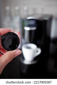 Close Up A Hand Holding A Coffee Capsule, With A White Coffee Cup And Coffee Machine On Black Counter, Blurred Background. 