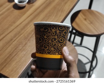 Close up of a hand holding a patterned brown and orange paper cup against a blurred cafe background. Disposable cup for coffee or tea service in a modern interior.