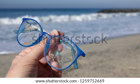 Close up of hand holding up a ruined, scratched and sandy pair of blue prescription glasses lost on beach                              