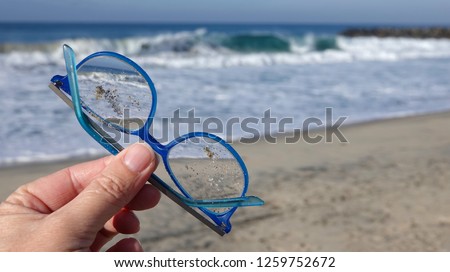 Close up of hand holding up a ruined, scratched and sandy pair of blue prescription glasses lost on beach                              