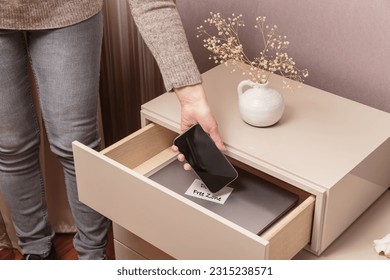 Close up of hand holding smartphone in separate desk drawer labeled Device Free Zone. Woman putting her phone in drawer with different gadgets at home. Digital detox and technology addiction concept