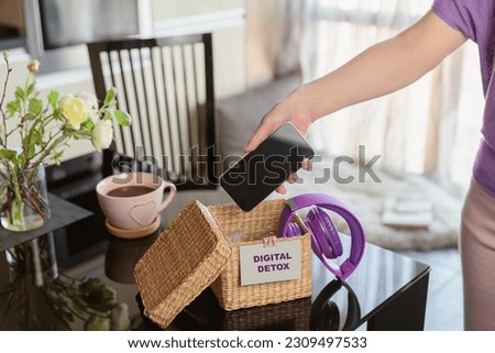Close up of hand holding smartphone in wicker basket on table. Woman putting phone into box with different gadgets at home. Digital detox and technology dependance concept