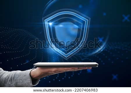 Close up of hand holding tablet with creative glowing shield hologram on blue background with various blurry icons. Cyber security, data protection concept. Modern wireframe design