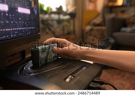 Close up of hand inserting arcade 8bit game cartridge into retro gaming console with TV displaying video game in cozy living room environment