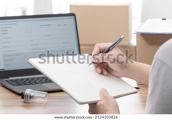 Close\
up hand a man hold pen write on notebook and car headlight bulb\
halogen on table, check detail of part for car service concept and\
use laptop and checklist in service office store\
