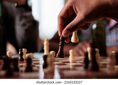 Close up of hand moving chess piece on chessboard while sitting at table. Person playing strategy boardgame with friends while sitting at home in living room with snacks and beverages.