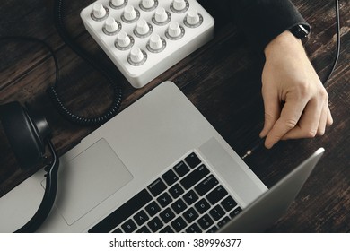 Close up hand plugs in headphone cord in stereo jack music port in retina laptop on vintage wooden table, side top view, midi mixer control and headphones near