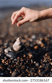 Close up of a hand stacking smooth stones on a pebble beach, forming a tiny zen cairn amid coastal rocks and waves, conveying calm balance and focus