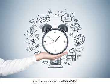 Close up of a hand of an unrecognizable businessman holding an alarm clock with a time management sketch around it near a gray wall.