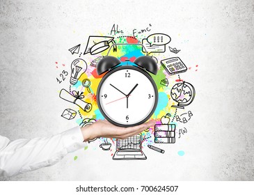 Close up of a hand of an unrecognizable businessman holding an alarm clock with a time management sketch around it near a concrete wall.