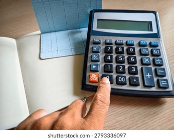 Close up of a hand using a calculator to record expenses in a bank passbook on a wooden desk, financial planning concept. 