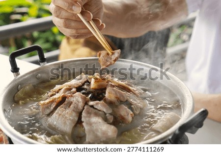 Close up of hand using chopsticks picking grilled pork from a traditional Thai BBQ pan (Mu Kratha) with hot steam and soup.