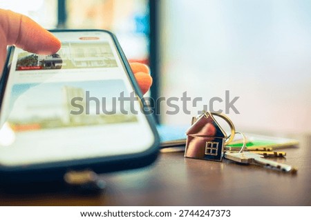 Close up of hand using smartphone searching for real estate house on screen with key on table. Digital property investment, online mortgage application, and mobile house hunting concept.