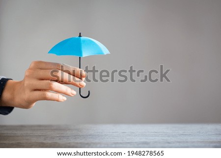 Close up hand of woman holding small umbrella on table with copy space. Woman holding small light blue umbrella as sign of protection on desk against grey wall. Girl protect something on table.