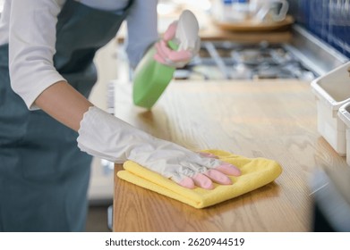 Close up hand of woman wearing rubber gloves cleaning wooden kitchen counter using clothes and cleaning solution. Home cleaning cooking hygienic wood surface care concept.