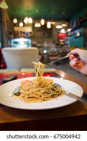 Close up hand of women holding fork with spaghetti with salted fish,  herbs and dried chilli  in thai style on wooden table with blurred of vintage restaurant background. Soft focus.