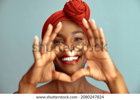 Close up of hands of beautiful african woman making heart shape isolated against blue wall. Portrait of smiling middle aged black woman making heart gesture with hands around her big white smile.