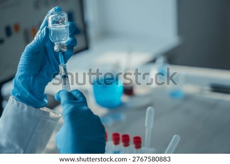 Close up of hands in blue gloves preparing an injection with a syringe and vial, surrounded by laboratory equipment. The image emphasizes medical research, vaccination, and scientific innovation