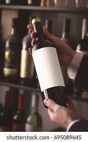 close up of hands of Caucasian men with white shirt holding a red wine bottle with white blank label against the sale shelves of a wine store or restaurant in natural light