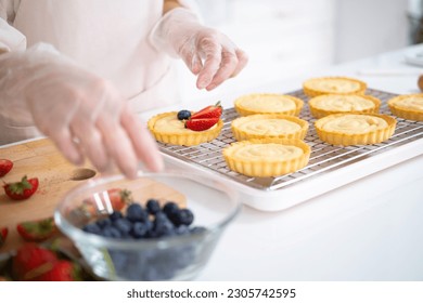 close up of hands in cooking gloves Baker adding blueberries strawberry fresh fruit to a tart on white table in Kitchen. housewife baker wear apron making fruit tart. homemade bakery at home. - Powered by Shutterstock