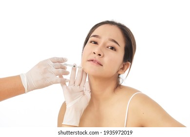 Close Up Of Hands Of Cosmetologist Making Botox Injection In Female Face. She Is Holding A Syringe. The Young Beautiful Asian Woman Is Receiving Procedure With Enjoyment