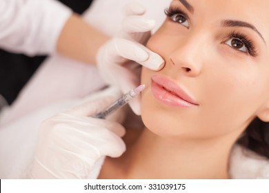 Close up of hands of cosmetologist making botox injection in female lips. She is holding syringe. The young beautiful woman is receiving procedure with enjoyment
