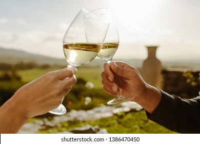 Close up of hands of a couple toasting glasses of white wine. Cropped shot of a couple toasting wine sitting in a wine farm.
