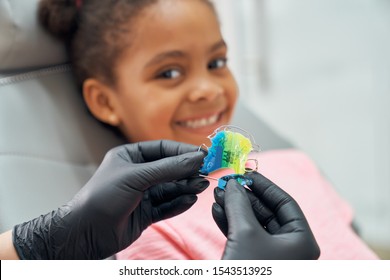 Close up of hands of dentist in black rubber gloves holding colorful braces. Pretty girl sitting in dental chair looking at camera, smiling. Applying braces for improving bite.
