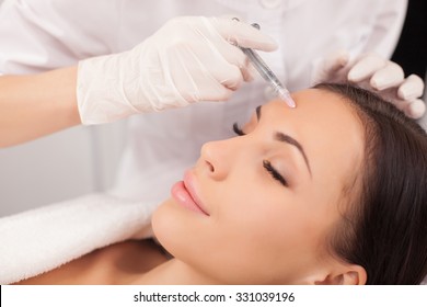 Close up of hands of expert beautician injecting botox in female forehead. The woman closed her eyes with joy. She is gently smiling