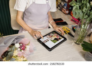 Close up hands of female wearing apron, working in flower shop. Pretty young female florist using tablet to chat with client, customer, scroll news feed. Social media, technology, business concept. - Powered by Shutterstock