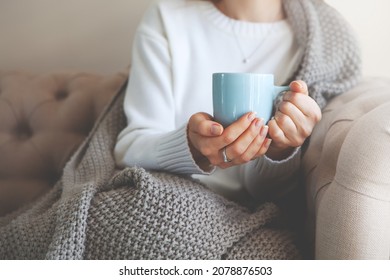 Close up of hands holding blue cup of tea or coffee. Model in white sweater and cozy plaid is sitting, relax at home on sofa. Depth of field, empty space on blurry beige background