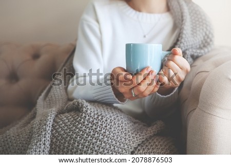 Close up of hands holding blue cup of tea or coffee. Model in white sweater and cozy plaid is sitting, relax at home on sofa. Depth of field, empty space on blurry beige background