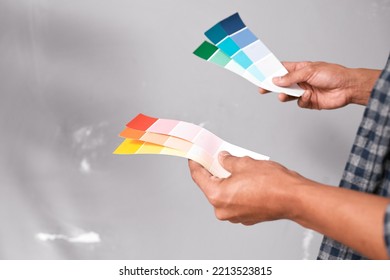 Close Up Of Hands Holding Bright Coloured Paint Swatches Against Grey Wall