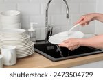 Close up hands of housewife washing dishes in at the black kitchen sink with white brush. Housework, cleaning plates with dishsoap,white subway tile backsplash