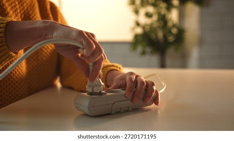 Close up of hands inserting a power plug into a surge protector on a table, with a blurred background.