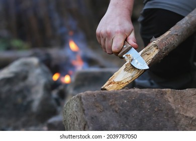 Close up of the hands of a man carving off timber to lit a fire, camp fire on the background. Hand holding knife cutting a wooden stick. Bushcraft and outdoors survival activities concept. 