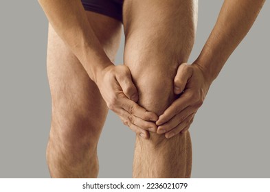 Close up of hands of man holding and massaging sore knee standing on gray background. Unknown man experiences joint pain while having arthritis or tendon problems. Health concept. Medical banner. - Shutterstock ID 2236021079