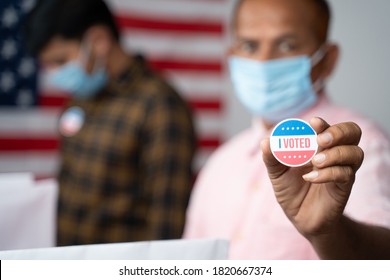 Close Up Of Hands, Man In Medical Mask Showing I Voted Sticker At Polling Booth With US Flag As Background - Concept In Person Voting At US Election