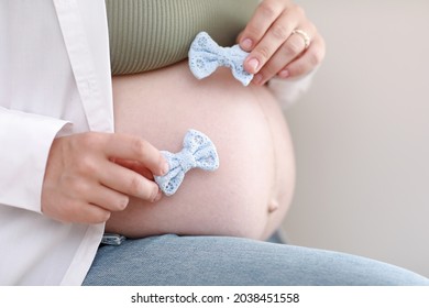 Close Up Hands On Pregnant Mother Holding Baby Clothes, Two Blue Bows. Lover Woman Waiting Child. Preparation For Childbirth, Girl Big Belly Advanced Pregnancy.