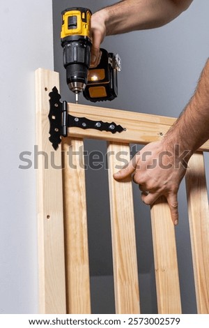 Close up of hands operating a yellow and black cordless drill to attach a black hinge to a light wooden gate with vertical slats indoors.
